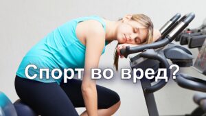 Изображение представляет собой цветную фотографию, изображающую обстановку спортзала, на которой изображена молодая женщина, отдыхающая на велотренажере. Изображение горизонтальное, на переднем и заднем плане изображено несколько тренажеров. На женщине со светлой кожей и прямыми каштановыми волосами, зачесанными назад, бирюзовый топ без рукавов и черные леггинсы. Она сидит на велосипеде, наклонившись вперед, положив голову на руль, с закрытыми глазами, что говорит об усталости или отдыхе. Современные велотренажеры оснащены цифровыми дисплеями и черно-серыми компонентами. На заднем плане - простая белая стена, что усиливает внимание к предмету и оборудованию. Общая атмосфера создает ощущение усталости или расслабления в фитнес-среде.